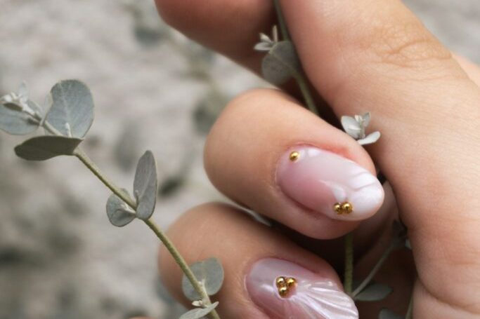 Gros plan d’ongles courts en nude laiteux au fondu discret, rehaussés de perles dorées : un dégradé ombré entre deux teintes voisines.